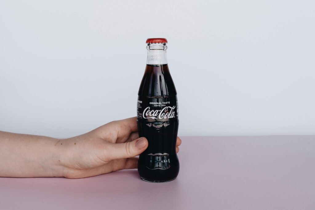 Close-up of a hand holding a classic Coca-Cola bottle on a pink surface, highlighting the refreshing drink.